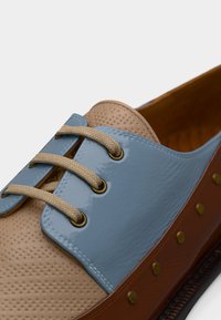 Καφέ και μπεζ παπούτσι με μπλε δερμάτινο επάνω μέρος, διάτρητη υφή, μπεζ κορδόνια και μεταλλικά καρφιά κατά μήκος της πλευράς. Λεπτό φινίρισμα, στρογγυλεμένο σχήμα.