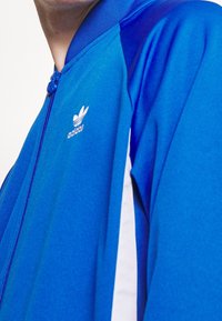 Sininen urheilutakki, jossa on vetoketjullinen kaulus, valkoiset sivuraidat ja pieni valkoinen Adidas-logo rinnassa. Sileä pinta.