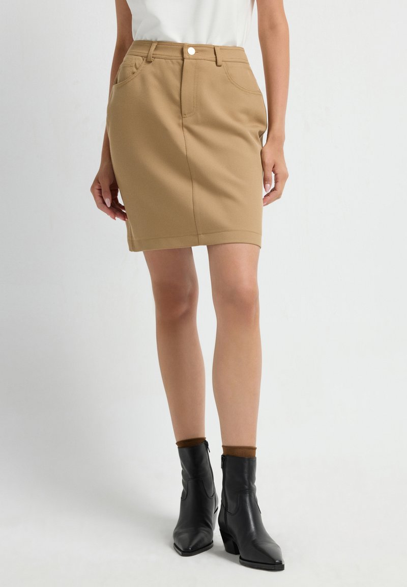 Khaki stretch denim rok met een aangesloten pasvorm, voorzakken en een tailleband met een zilveren knoopaccent. G gedragen met zwarte enkelboots.