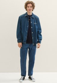 Giacca in denim e pantaloni di un blu medium wash. La giacca ha chiusure a scatto e polsini elasticizzati. Sneakers nere con suole bianche.