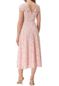 Femme portant une robe midi en dentelle rose avec des manches courtes, un dos en V, et des sandales à talons hauts couleur nude, debout de dos.