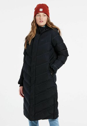 Manteau d'hiver long noir avec un motif en chevrons, col haut et fermetures éclair sur les poches latérales. Porté avec un bonnet en tricot rouge.