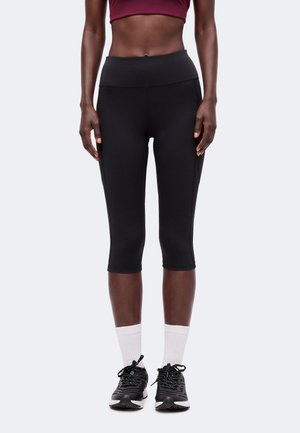 Personne athlétique portant des leggings courts noirs, des chaussettes blanches, des chaussures de course noires et un soutien-gorge de sport rouge foncé, debout devant un fond clair.
