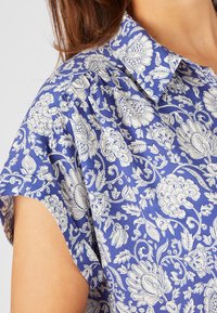 Blusa floral azul con patrones botánicos blancos, cuello puntiagudo, mangas cortas y textura suave y lisa.