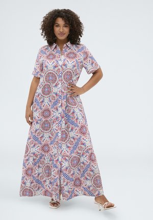 Mujer de pie con un vestido de manga corta hasta el tobillo con intrincados patrones florales y de cachemira en rojo, blanco y azul.