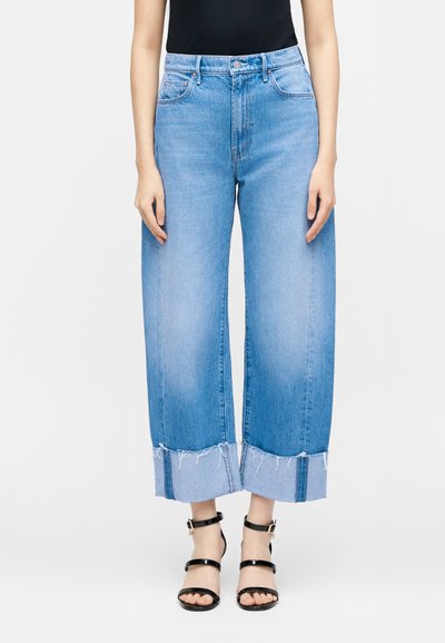 Γυναίκα που φοράει ανοιχτό μπλε τζιν παντελόνι με φαρδιά μπατζάκια και ξεφτισμένα τελειώματα, καθώς και μαύρα πέδιλα με λουράκια και ψηλά τακούνια, στέκεται μπροστά σε λευκό φόντο.