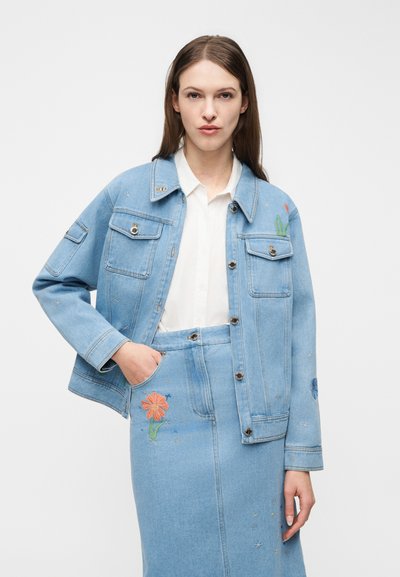 Kvinde iført lyseblå denimjakke og nederdel med farverig blomsterbroderi over en hvid skjorte med knapper, stående med hånden i lommen.