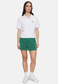 Polo shirt bianco in cotone con maniche corte e colletto, abbinato a pantaloni corti in cotone verdi. Logo Fila ben visibile sulla maglietta e sui pantaloni.
