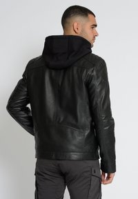 Schwarze Lederjacke mit abnehmbarer schwarzer Kapuze. Gepolsterte Schulterdetails. Glatte Textur, taillierter Schnitt und keine sichtbaren Verschlüsse auf dem Rücken.