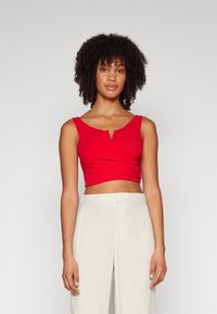 Top corto rosso con scollatura a V e design frontale annodato, realizzato in un tessuto liscio. Abbinato a pantaloni a vita alta di colore chiaro.