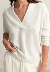 Pull en maille blanc, doux, avec un décolleté en V profond et des manches longues, offrant une coupe décontractée. Assorti à un pantalon blanc taille haute.