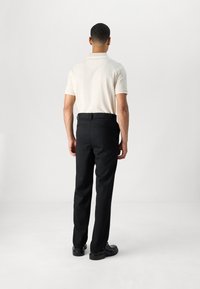 Beige Poloshirt mit kurzen Ärmeln, kombiniert mit schwarzen Hosen, Ansicht von hinten. Das Material wirkt glatt, mit klassischer Passform und ohne auffällige Details.