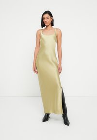 Μάξι φόρεμα από σατέν σε ελιά πράσινο, με τετράγωνη λαιμόκοψη και λεπτές τιράντες. Φοριέται με μαύρες, ψηλές μέχρι το γόνατο, μυτερές μπότες. Ομαλή υφή.