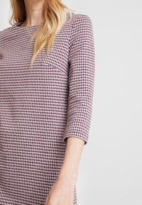 Femme portant une robe cintrée avec un motif pied-de-poule rouge, blanc et noir et des manches trois-quarts sur un fond uni.