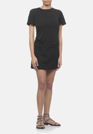 Donna in abito nero a maniche corte e sandali neri con cinturino alla caviglia e borchie, su sfondo semplice.