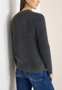 Šedý chlupatý svetr s kulatým výstřihem a dlouhými rukávy, vyrobený z měkkého texturovaného materiálu. Kombinován s modrými džínami. Jednoduchý, minimalistický design.