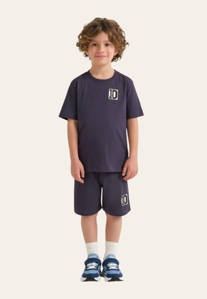 Bambino piccolo con capelli ricci che indossa un completo con t-shirt e pantaloncini blu navy con loghi, calzini bianchi e scarpe da ginnastica blu, in piedi con il viso rivolto in avanti.