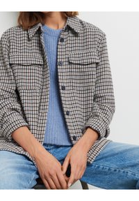 Femme portant un pull en laine bleu sous une veste boutonnée marron et noire à carreaux, associée à un jean bleu clair, les mains reposant sur les genoux.