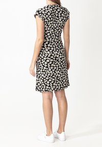 Kobieta w czarnej sukience do kolan z białym kwiatowym wzorem i białych trampkach, stojąca na tle jednolitego białego tła.