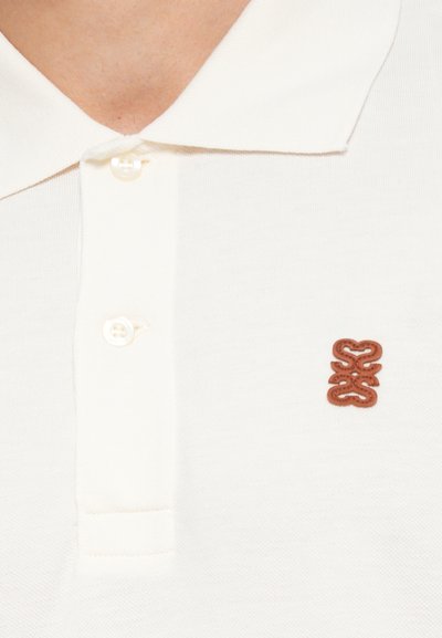 Hvid polo-shirt lavet af tekstureret stof med en to-knap lukning. Har et brunt broderet logo på venstre side over knapperne.