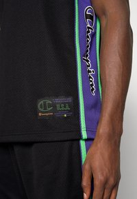 Černé síťované sportovní šortky s fialovými a zelenými bočními pruhy a nášivkou loga Champion, modelova ruka viditelná na pravé straně.