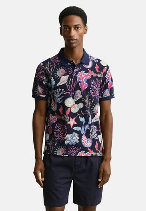 Mann trägt ein marineblaues Poloshirt mit bunten Muschel- und Korallenmustern, kombiniert mit dunkelblauen Shorts, steht vor einem schlichten Hintergrund.