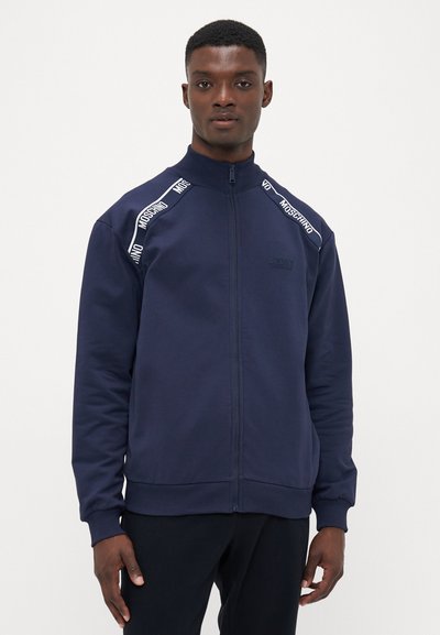 Sweat-shirt zippé marine avec col haut, accents en bande blanche "MOSCHINO" sur les épaules, poignets côtelés et texture de tissu lisse.
