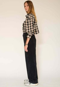 Chemise à carreaux noire et crème avec des volants, associée à un pantalon en velours côtelé foncé et des baskets argentées. Le mannequin se tient de côté, montrant la tenue.