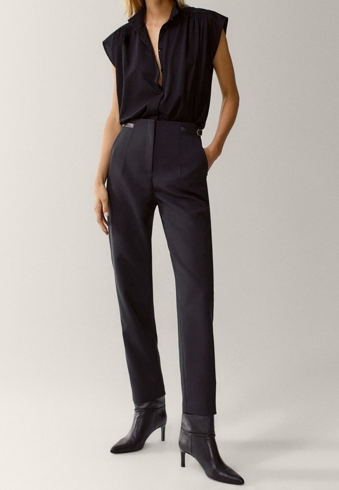 Femme portant un chemisier noir sans manches, un pantalon noir taille haute et cintré, et des bottines noires à talons pointus, debout avec une main dans la poche.