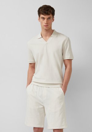 Młody mężczyzna z krótkimi kręconymi włosami, ubrany w kremową koszulkę polo z krótkim rękawem i pasujące krótkie spodenki, stoi z rękami w kieszeniach.
