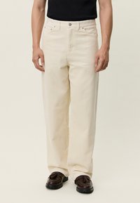 Bež kordurove hlače ravnog kroja, s prednjim džepovima i zatvaranjem na gumbe, u kombinaciji s tamno smeđim cipelama. Teksturirani materijal s uspravnim prugama.