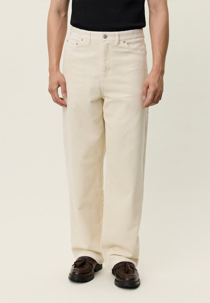 Bež kordurove hlače ravnog kroja, s prednjim džepovima i zatvaranjem na gumbe, u kombinaciji s tamno smeđim cipelama. Teksturirani materijal s uspravnim prugama.