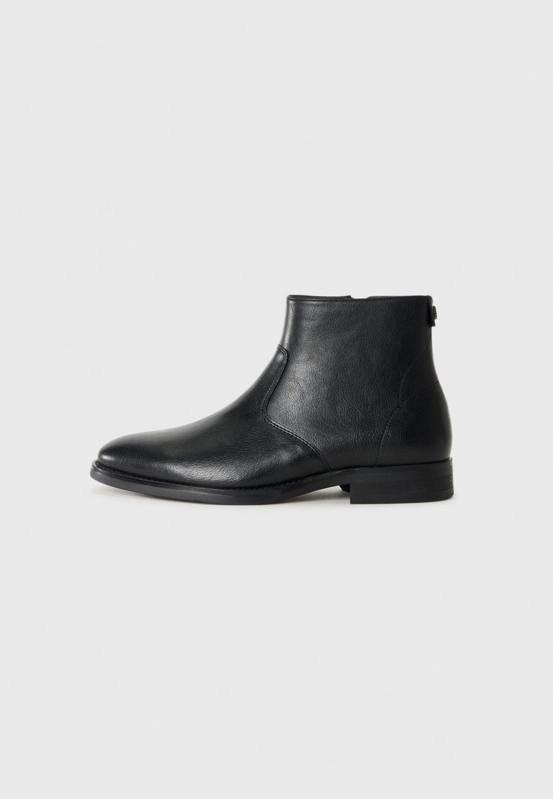 Bota de tobillo negra de cuero con punta redonda, textura suave, tacón bajo y detalle de cremallera lateral para facilitar su uso.