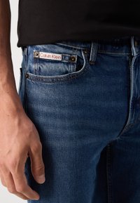 Kék farmernadrág enyhén kopott felülettel, látható Calvin Klein logóval egy fehér címkén, és fém kiegészítőkkel a zsebeken.