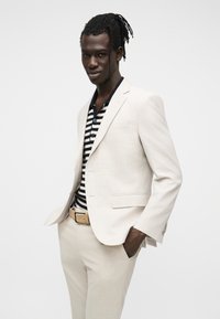 Homme avec dreadlocks portant un costume beige, une chemise rayée noire et blanche, et une ceinture beige, debout les mains dans les poches sur un fond blanc.