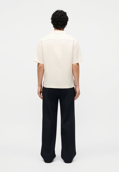 Άνδρας με σγουρά μαλλιά, που φοράει μπεζ κοντομάνικο πουκάμισο και φαρδιά μαύρη παντελόνα, στέκεται με την πλάτη προς το μέρος μας σε απλό φόντο.