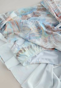 Tissu plié avec un motif pastel de coquillages en tons bleu clair, beige et rose, montrant la taille d'un vêtement et une texture douce.