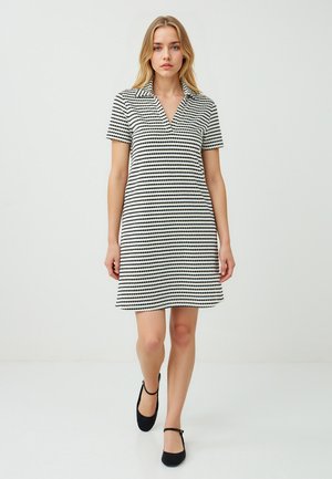 Una donna con lunghi capelli indossa un vestito corto con colletto, caratterizzato da righe bianche e nere, camminando con sicurezza su uno sfondo chiaro.
