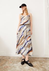 Maxi-jurk met een V-hals, voorzien van een draaiend, multikleurig marmerpatroon in tinten blauw, oranje en beige; gecombineerd met zwarte schoenen en een pet.