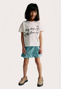 Camiseta blanca de manga corta con texto y diseños bordados en negro, combinada con una falda vaquera azul claro con volantes y zapatos beige.