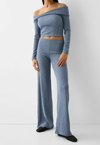 Lichtblauwe, geribbelde gebreide off-shoulder crop top, gecombineerd met bijpassende wijde broek. De stof is textuurrijk en zacht, met lange mouwen.
