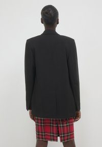 Schwarzes, maßgeschneidertes Blazer mit strukturierten Schultern, getragen über einem roten Karorock, zeigt einen geraden Rücken und eine schlanke Passform. Texturiert.