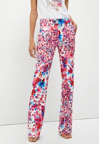 Vrouw in witte broek met een levendig rood, roze en blauw bloemenpatroon, gecombineerd met een witte top en oranje open teen sandalen.