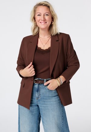 Blonde vrouw die glimlacht, een bruine blazer en top draagt, blauwe spijkerbroek, gouden creolen als oorbellen, gelaagde kettingen en meerdere armbanden aan haar rechterpols.