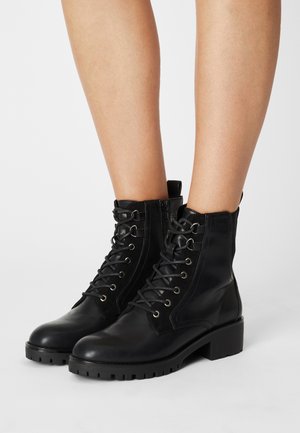 Bottes noires en cuir à lacets montantes avec fermetures éclair latérales et talons épais portées sur jambes nues sur fond blanc.