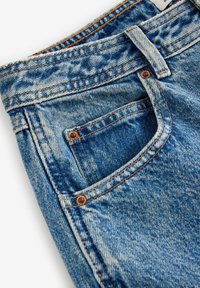 Gros plan sur un jean en denim bleu montrant la ceinture, la passants, la poche avant avec rivets et les coutures orange détaillées.