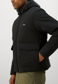 Homme portant une veste matelassée noire avec capuche et poches avant, associée à un pantalon vert olive, se tenant devant un fond uni.