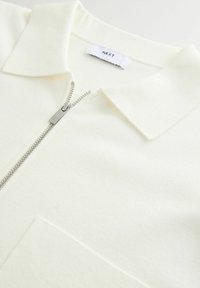 Hvid zip-up polo med en blød tekstur, der har en klassisk krave og en frontlomme. Sølvfarvet lynlås hardware tilføjer en subtil accent.