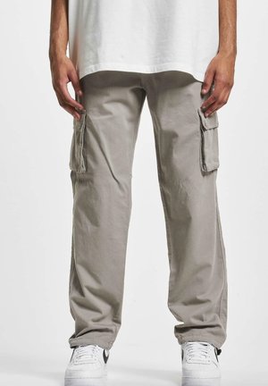 Man met beige cargobroek en witte sneakers, staand met ontspannen handen langs het lichaam tegen een effen achtergrond.