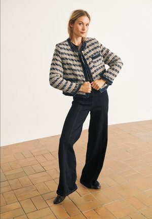 Femme portant une veste rayée texturée avec de gros boutons et un pantalon noir à jambes larges, debout sur un sol carrelé contre un mur blanc.